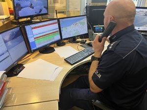 Dyżurny kołobrzeskiej jednostki na stanowisku kierowania - umundurowany policjant z słuchawką telefonu w ręku, stół z czterema monitorami