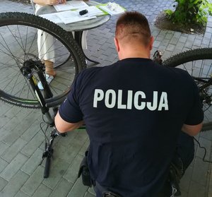 umundurowany policjant przy rowerze, kobieta przy biurku w tle