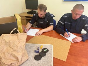 Dwóch umundurowanych policjantów piszących protokoły przy stole. Na stole torba papierowa, 3 czarne podstawki, 3 cukierki.