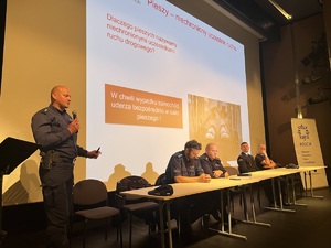 scena, na scenie przedstawiciele policji i straży pożarnej, z tyłu pulpit z tłem debata społeczna i logo policji kołobrzeg