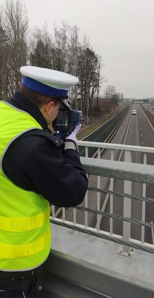 umundurowany policjant ruchu drogowego trzymający urządzenie do pomiaru prędkości stojący na wiadukcie, w oddali droga