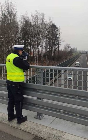umundurowany policjant ruchu drogowego trzymający urządzenie do pomiaru prędkości stojący na wiadukcie, w oddali droga